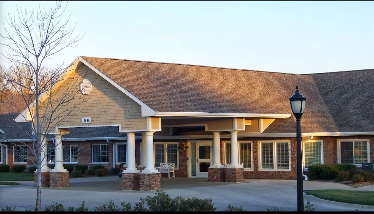 Exterior front view of a single-story senior living facility building with a covered entrance supported by white columns with stone bases. The building has a brown shingled roof, beige siding, and multiple windows with white frames. A black street lamp is visible in the foreground, and there are some leafless trees and shrubs around the building.