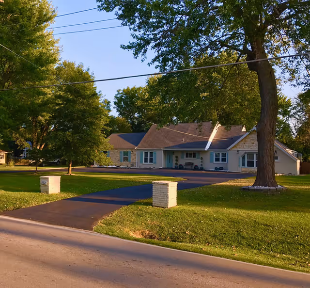 Single-story senior living building with a paved walkway, lawn, and large trees in front.