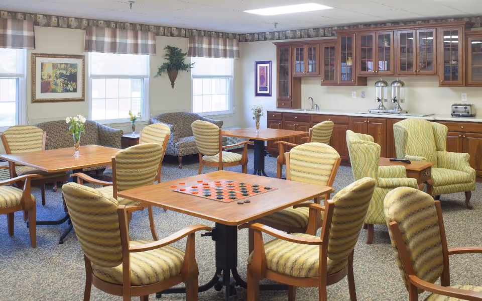 A well-lit common area with multiple wooden tables and cushioned chairs arranged around them. One table has a checkers board set up. The room features a kitchenette with wooden cabinets, a sink, and beverage dispensers. There are two large windows with valances, framed artwork on the walls, and two green upholstered armchairs with a small wooden side table between them.