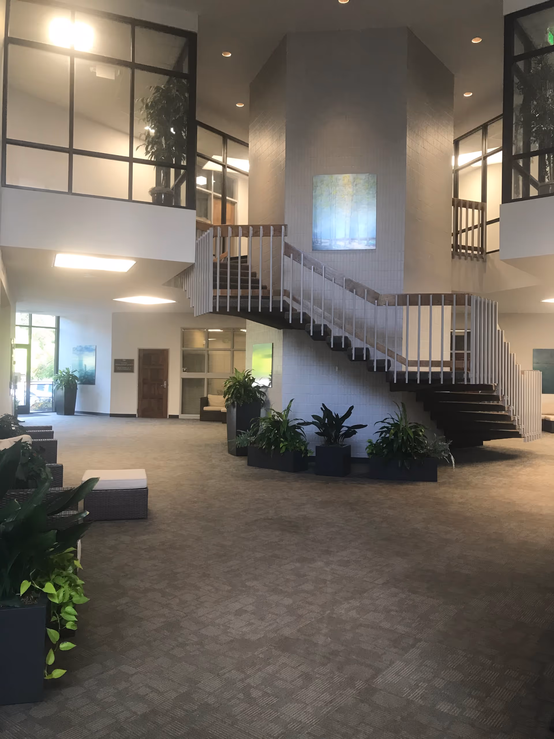 Bright lobby with central staircase, potted plants, and seating in a spacious facility interior.