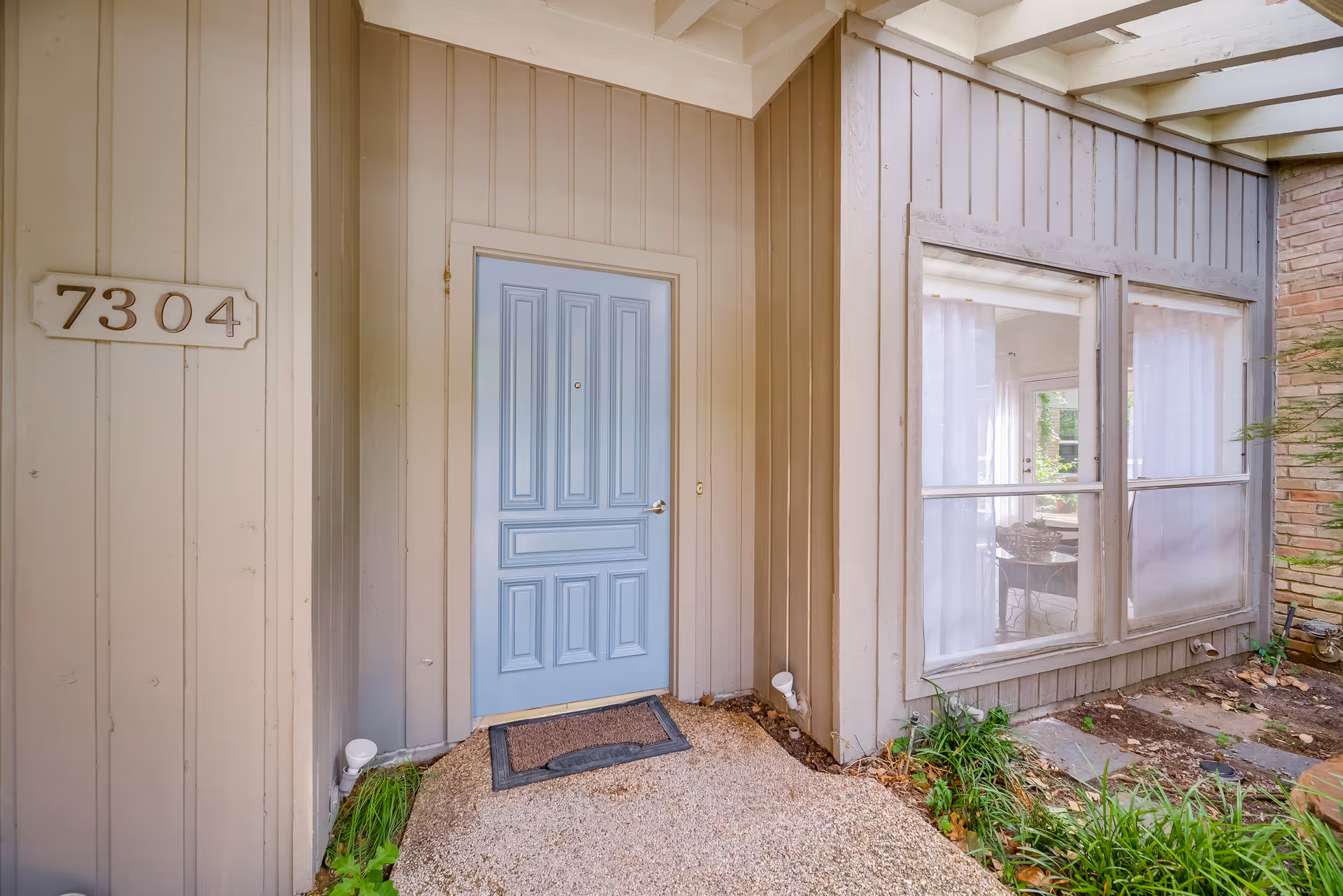 Exterior entrance of a home with a light blue front door, house number 7304 on the siding, and adjacent large windows.
