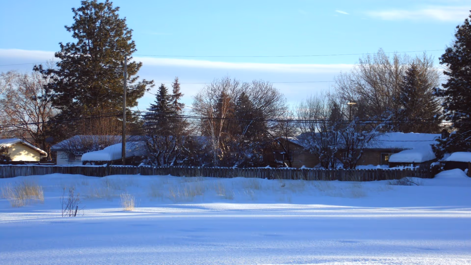 Snow-covered landscape with a wooden fence, trees, and houses in the distance.