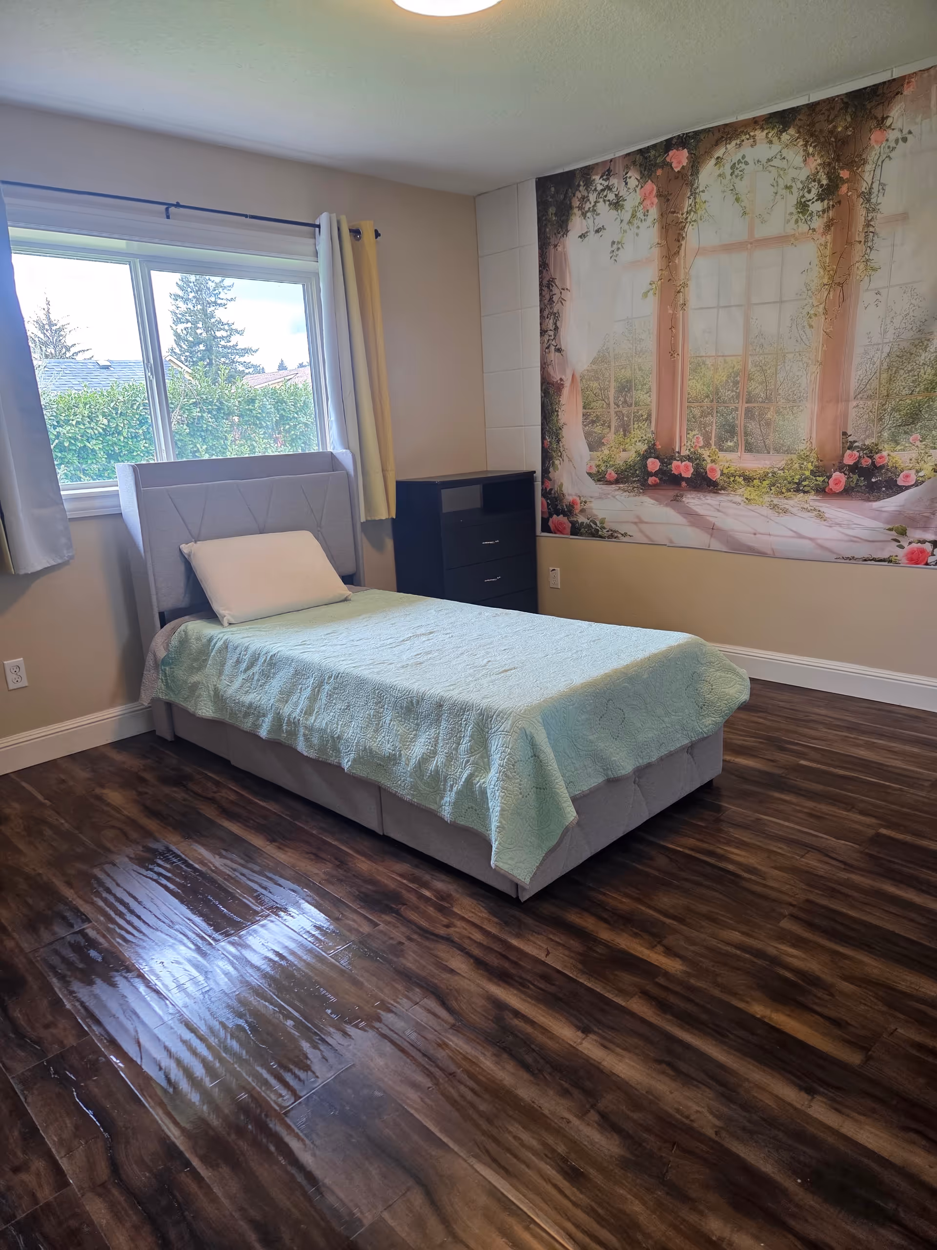 Bright bedroom in a senior living facility featuring a bed, dresser, window, and mural wall.