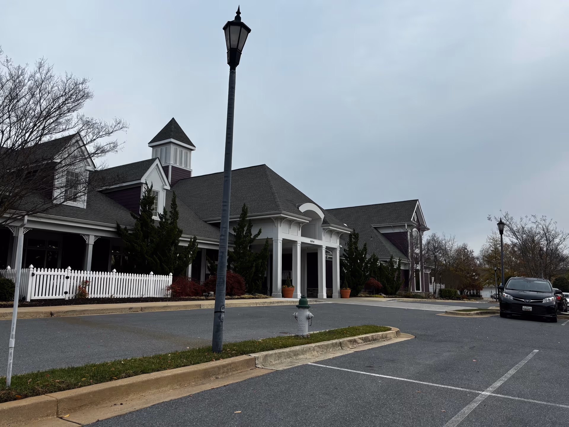 Exterior view of a senior living facility building with a peaked roof and white columns at the entrance. There is a white picket fence, some shrubs, a street lamp, a fire hydrant, and a few parked cars in the parking lot under an overcast sky.