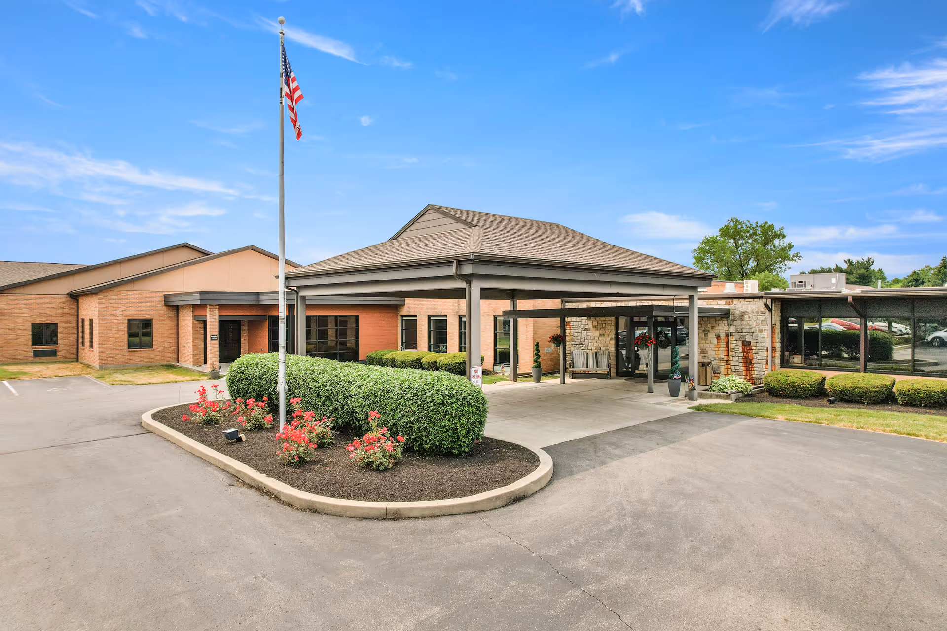 Exterior view of Vancrest of Urbana senior living facility showing the entrance with a covered drop-off area, an American flag on a flagpole, neatly trimmed bushes, and a clear blue sky.