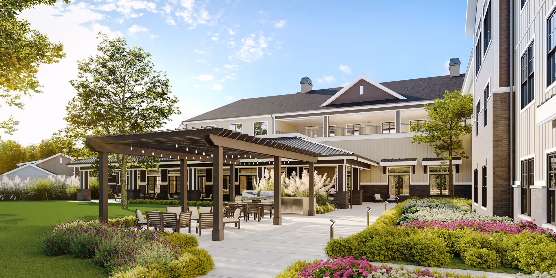 Outdoor patio area at a senior living facility with a pergola covering seating and dining tables. The area is surrounded by well-maintained landscaping including green bushes, purple and pink flowers, and trees. The building in the background is a multi-story structure with balconies and large windows.