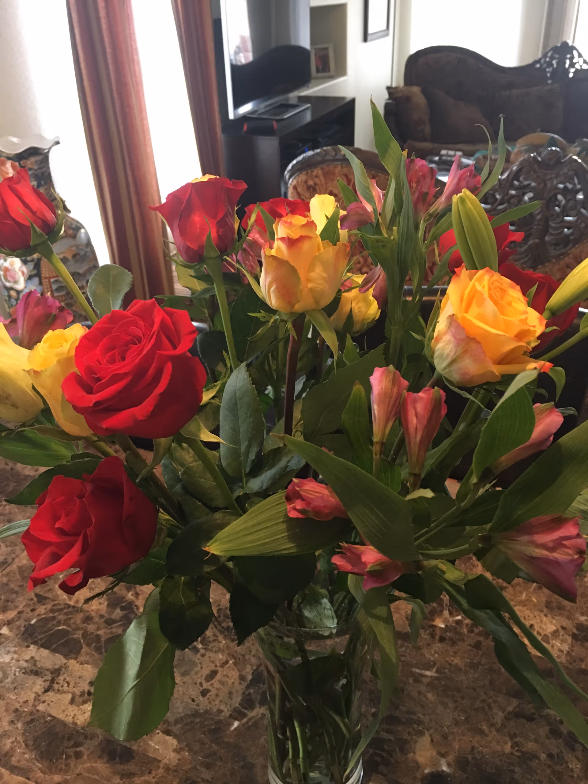 A bouquet of red and yellow roses with pink flowers and green leaves in a glass vase placed on a marble table. In the background, there is a living room area with a TV, wooden chairs, and a sofa.