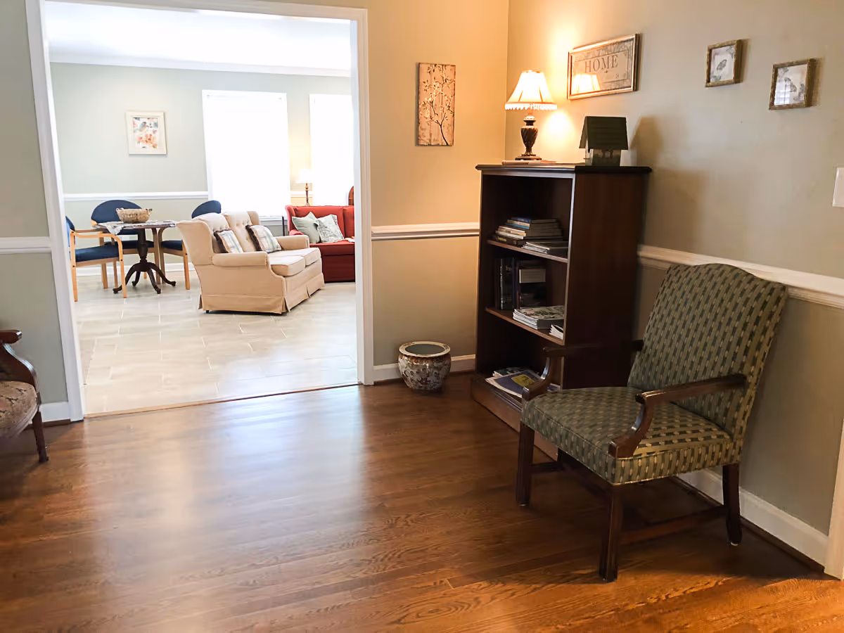 Interior view of a senior living facility showing a hallway with wooden flooring, a green patterned armchair, a wooden bookshelf with books and a lamp on top, and framed artwork on the beige walls. Through an open doorway, a living room area is visible with a beige loveseat, a red sofa, a small round table with chairs, and large windows letting in natural light.