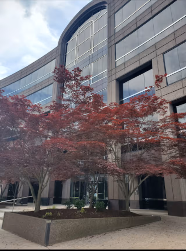 Modern glass-fronted senior living building with red-leaved trees and a paved entrance.