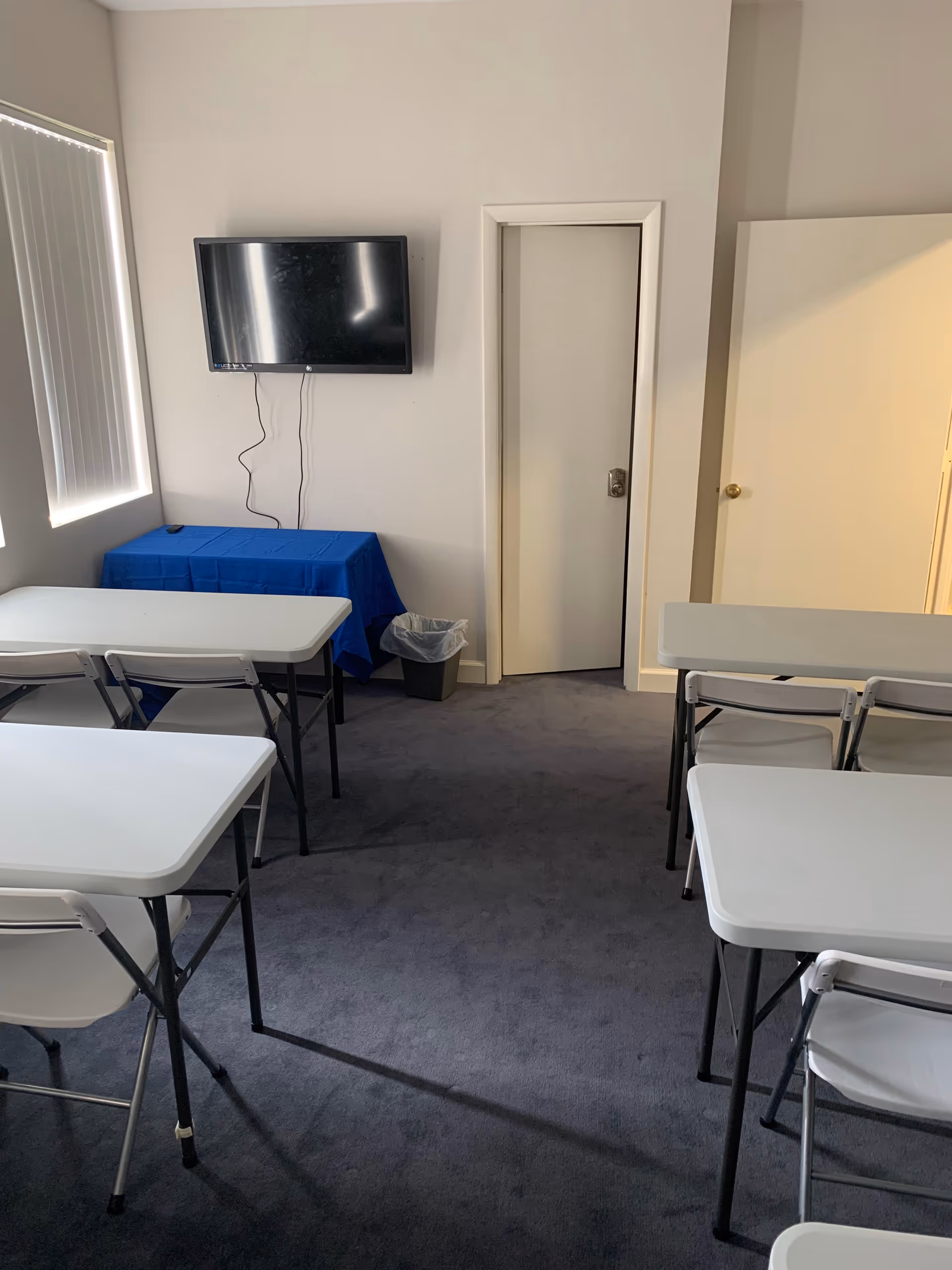 A small classroom-style room with white desks, chairs, a wall-mounted TV, and a door.