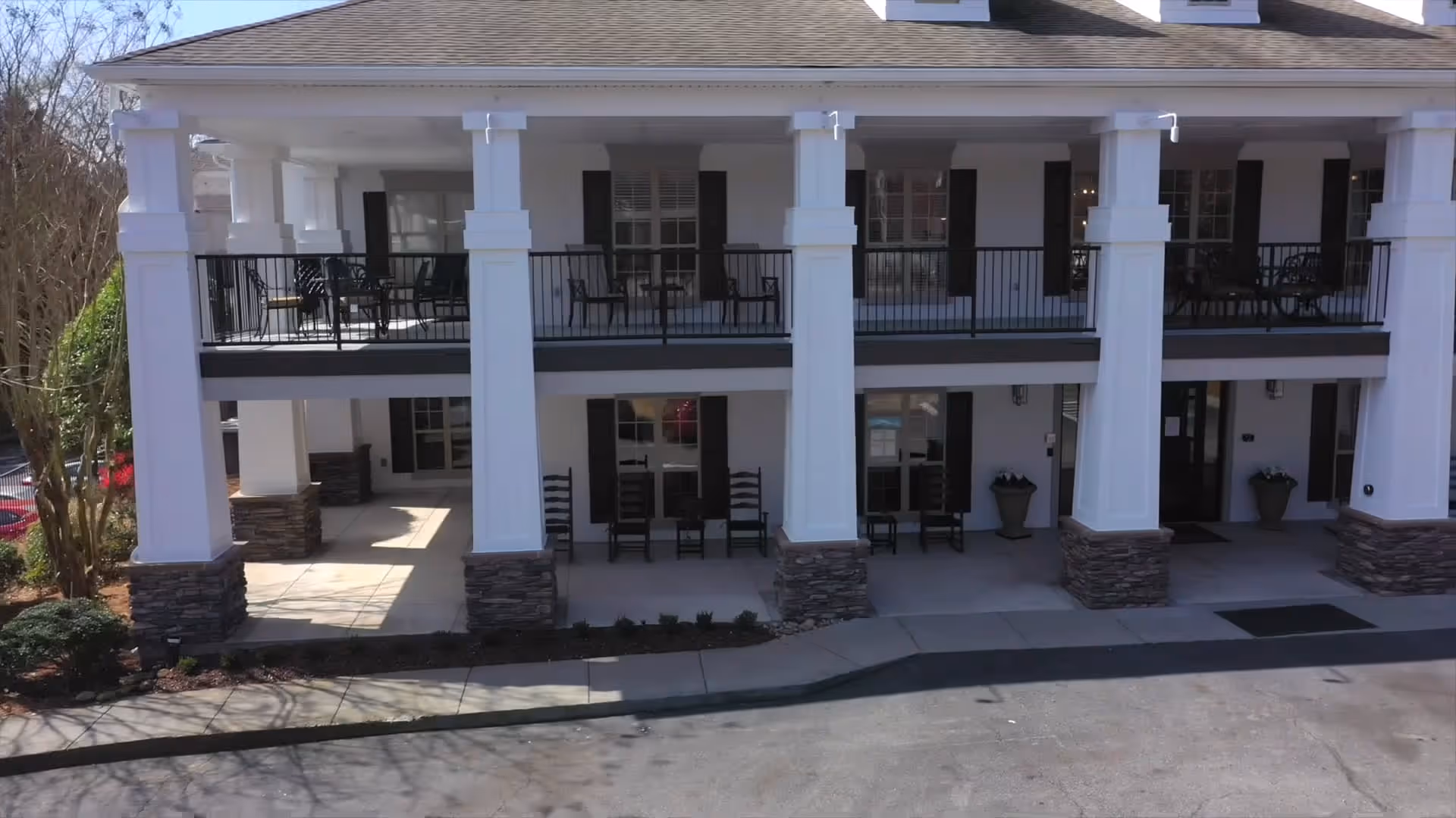 Front exterior view of a two-story building with white columns and stone bases. The upper level has a balcony with black railings and several chairs. The lower level has a covered porch area with rocking chairs and potted plants near the entrance.