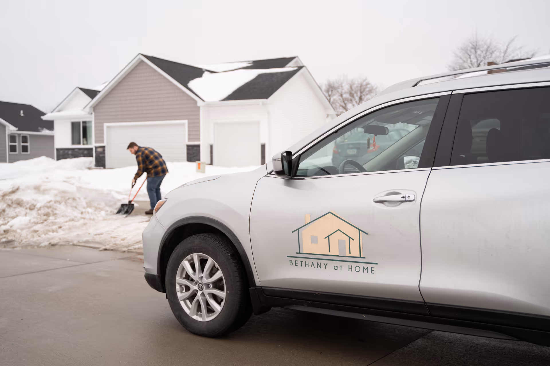 A Bethany at Home SUV parked outside a snowy residential neighborhood with a man shoveling snow.