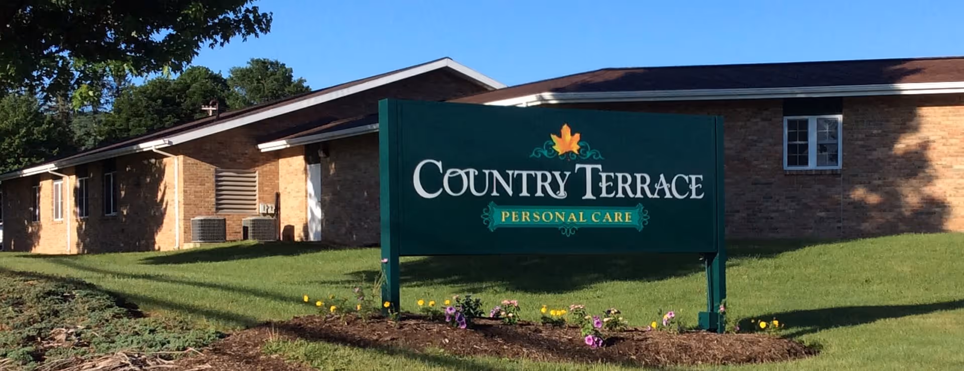 Exterior view of a single-story brick building with a green lawn and a sign in front that reads 'Country Terrace Personal Care' with a decorative leaf above the text.