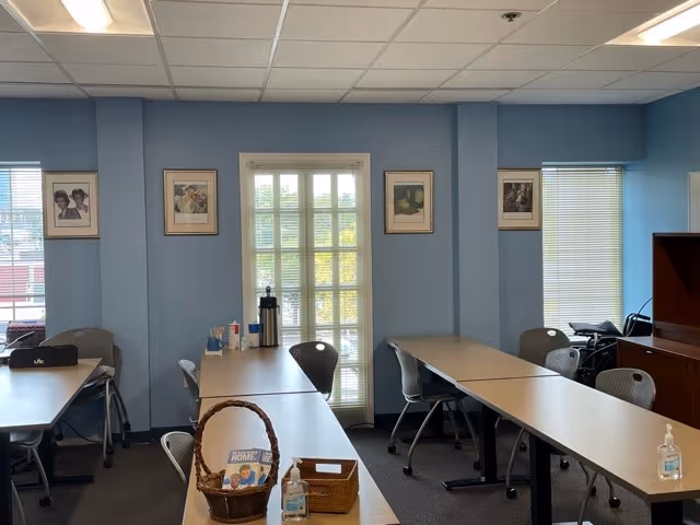 Light blue interior room with long tables, chairs, a doorway, and framed photos on walls.