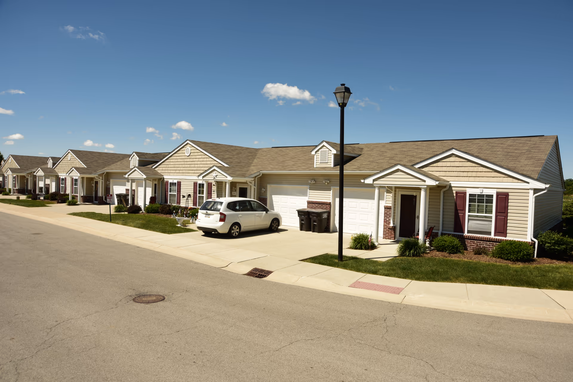 Row of beige townhome-style senior living units along a sunny street with a parked car.
