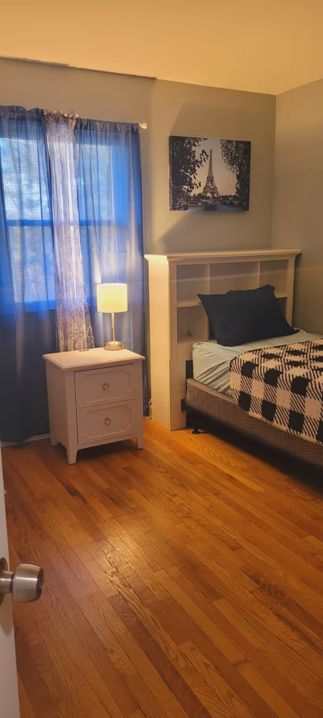 A cozy bedroom with wooden flooring, a bed with a black and white checkered blanket and a black pillow, a white nightstand with two drawers, a lit table lamp, blue curtains covering a window, and a wall art featuring the Eiffel Tower.