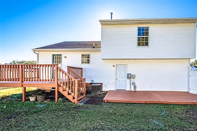 Exterior view of a two-story white house with a wooden deck and stairs leading down to a grassy yard under a clear blue sky.