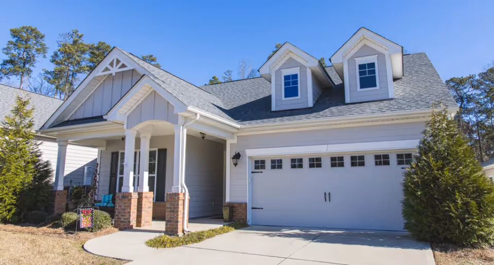 Exterior view of a single-story residential building with a gray roof, white siding, and brick accents. The house features a covered front porch with white columns and a two-car garage with decorative windows. There are trees and shrubs around the house under a clear blue sky.