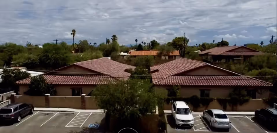 A view of single-story stucco buildings with red tile roofs and a parking lot with several cars surrounded by desert landscaping.