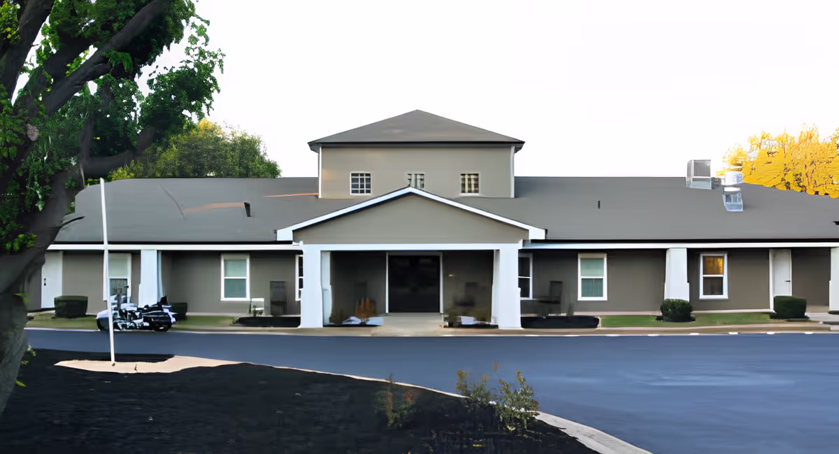 Front exterior view of a single-story senior living facility building with a central entrance covered by a small portico. The building is painted gray with white trim and has several windows. There is a paved driveway in front and some landscaping with trees and bushes around the building.