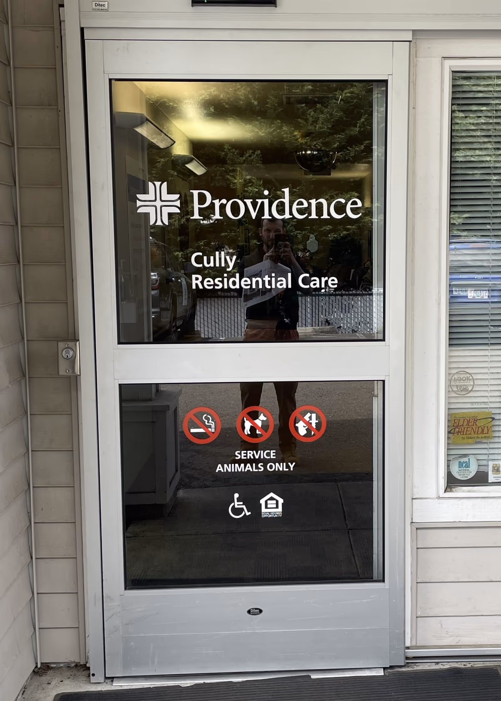 Glass entrance door displays Providence Cully Residential Care signage; reflection of person taking photo.