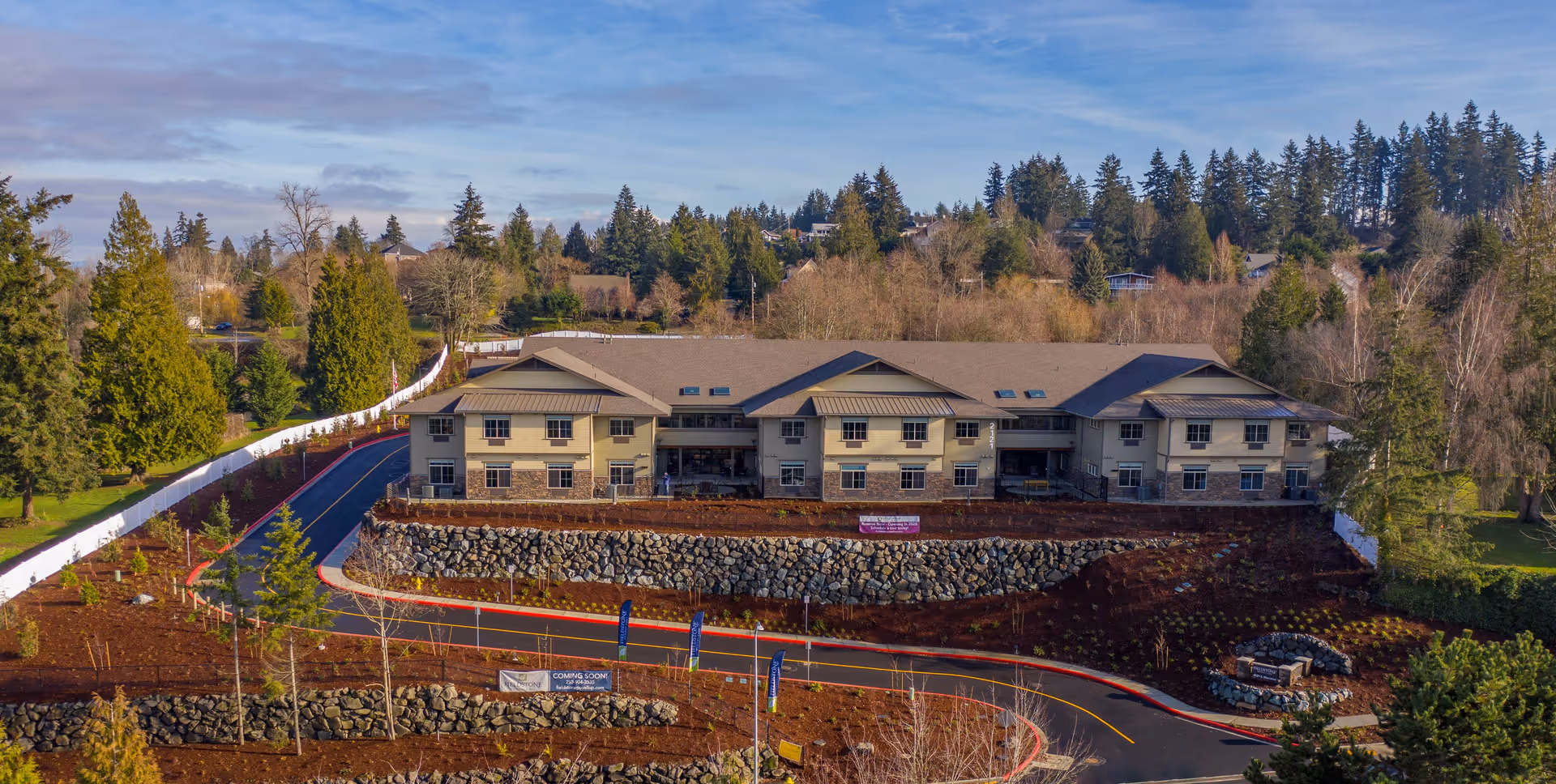 Aerial view of a two-story memory care building on a terraced, landscaped lot with surrounding trees and a winding driveway.