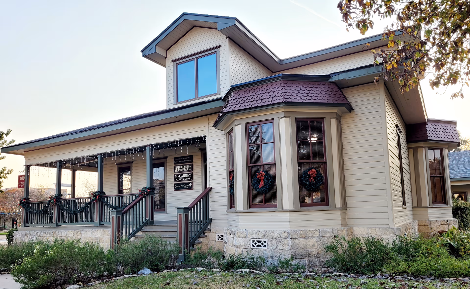 Exterior view of a beige building with a wraparound porch and holiday wreaths.
