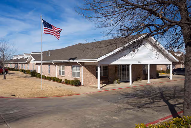 Single-story brick senior living building with a covered entrance, an American flag on a flagpole, and a driveway.
