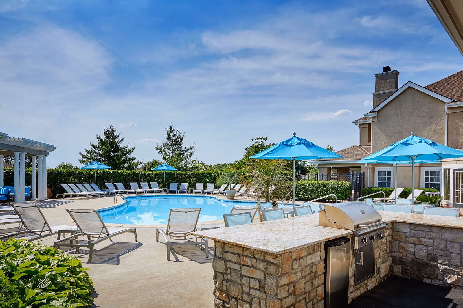 Outdoor pool area with lounge chairs, blue umbrellas, and a stone grill counter.