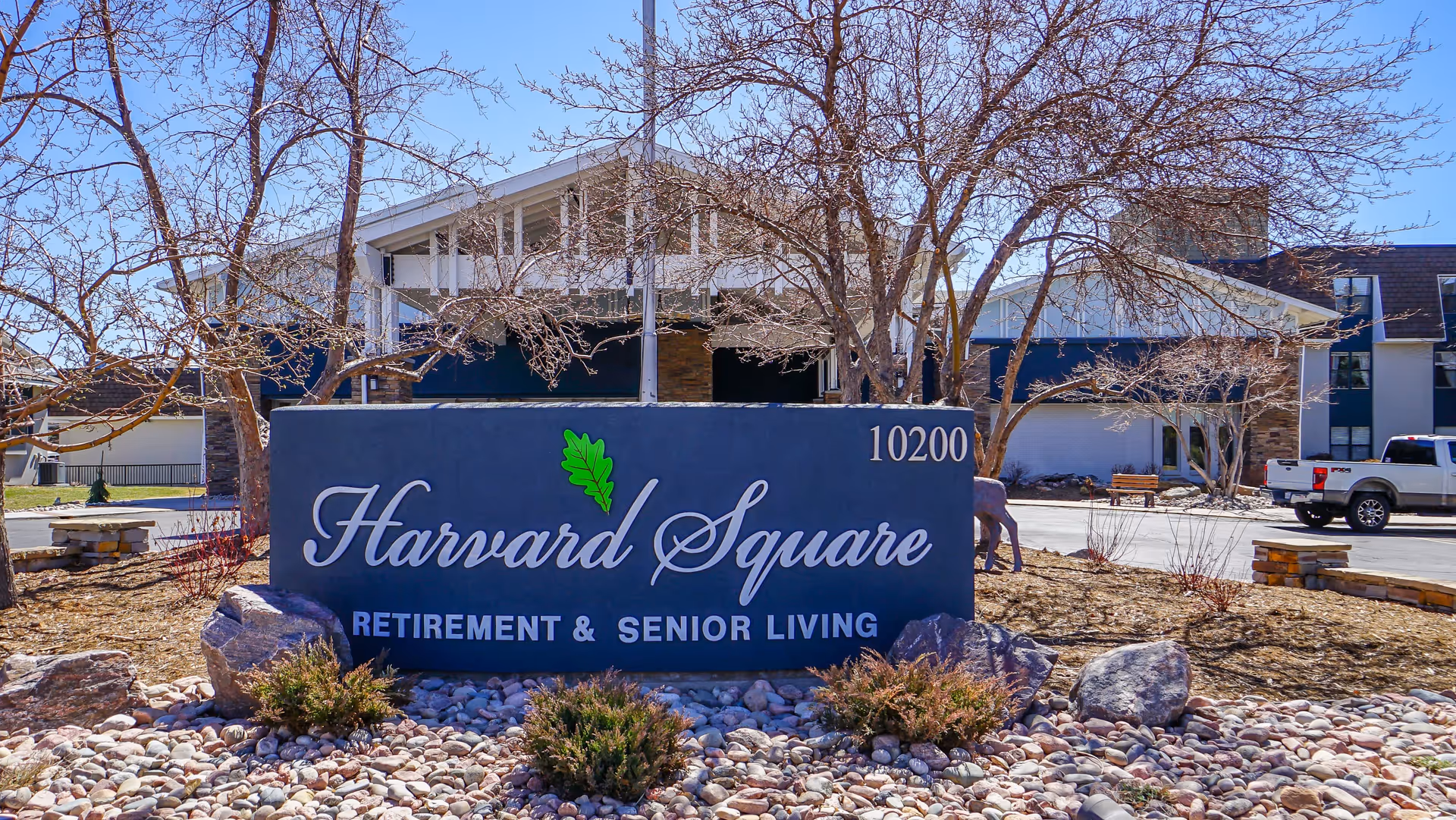 Exterior view of Harvard Square Retirement & Senior Living facility with a large sign in front displaying the name and address 10200. The building is surrounded by leafless trees and landscaping with rocks and bushes under a clear blue sky.