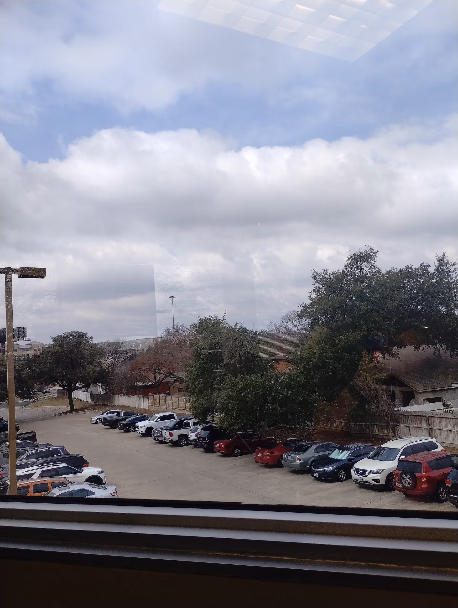 View from inside window of a parking lot with cars, trees, and cloudy sky.