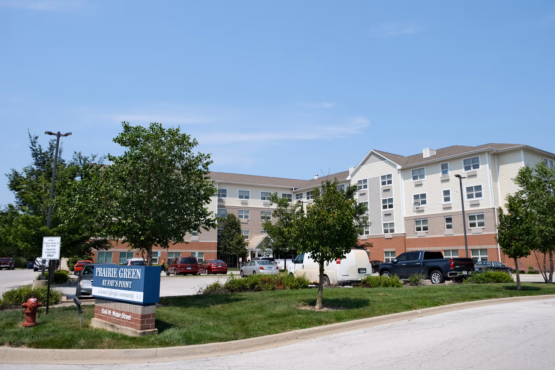 Front exterior of a multi-story senior living building with a sign reading "Prairie Green at Fay's Point" and parked cars in the lot.