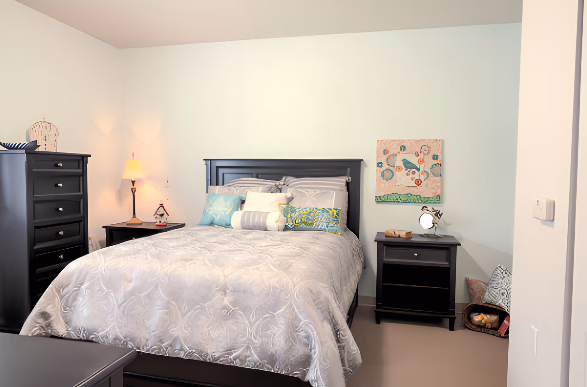A neatly made bed with a gray patterned comforter and multiple decorative pillows in a bedroom. The room features black wooden furniture including a tall dresser on the left and two nightstands on either side of the bed. A small lamp is lit on the left nightstand, and a colorful abstract painting hangs on the wall above the right nightstand. The walls are painted light blue, and there are some cushions and a basket on the floor near the right nightstand.