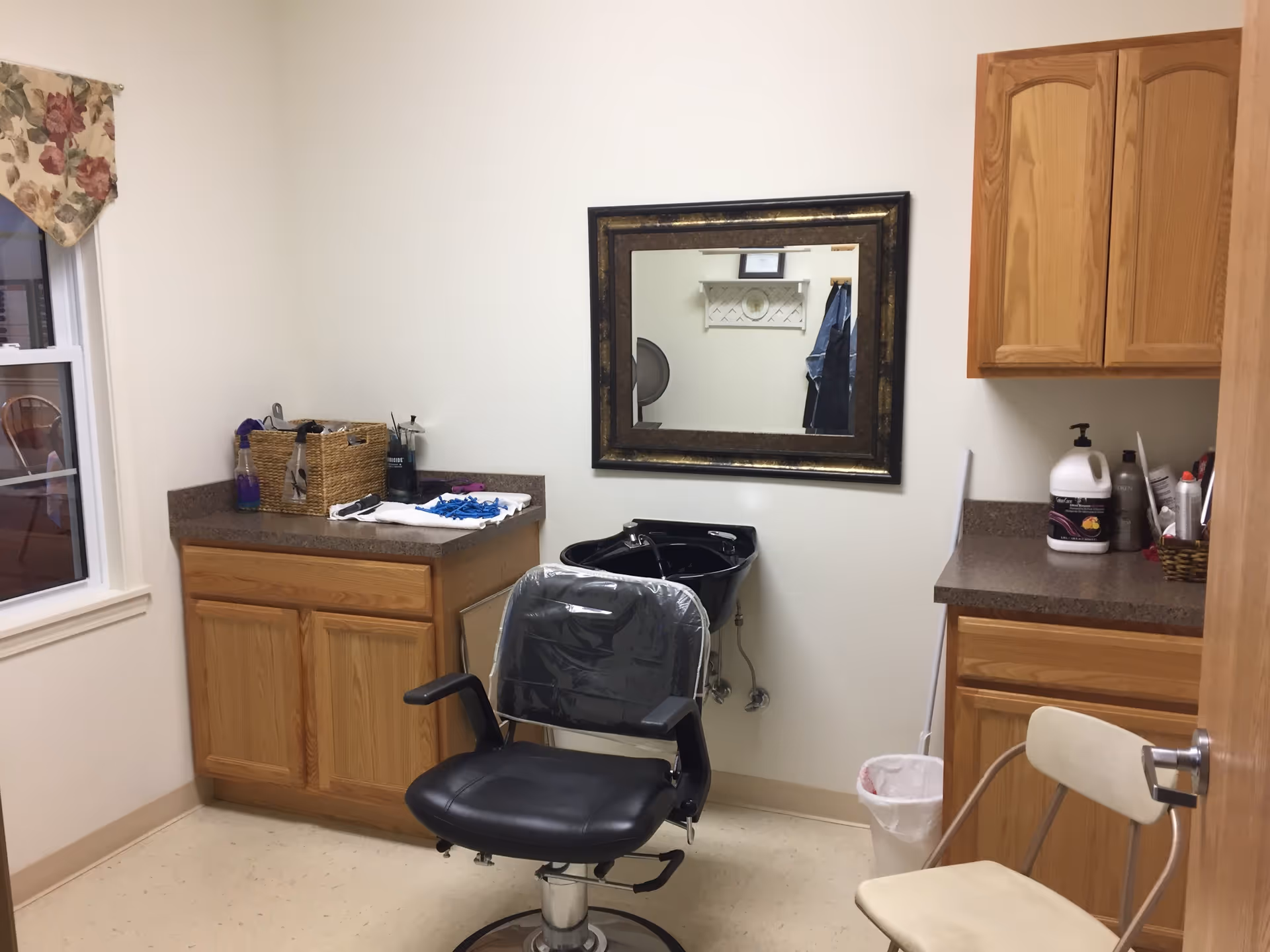 Interior of a small hair salon or barber station with a black salon chair covered in plastic, a black wash basin, a large framed mirror on the wall, wooden cabinets and countertops with various hair care products, and a window with floral curtains.