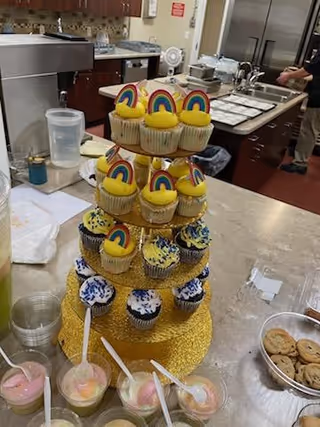 Tiered rainbow cupcake display on a glittery stand, set on a kitchen counter with cookies nearby.