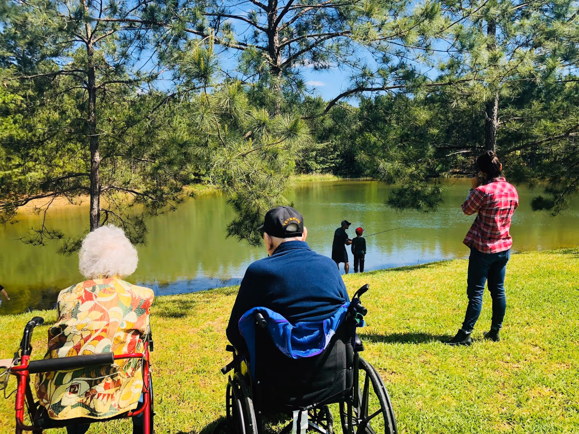 Two elderly residents in wheelchairs sit on a grassy bank overlooking a tree-lined pond while others stand nearby and fish.