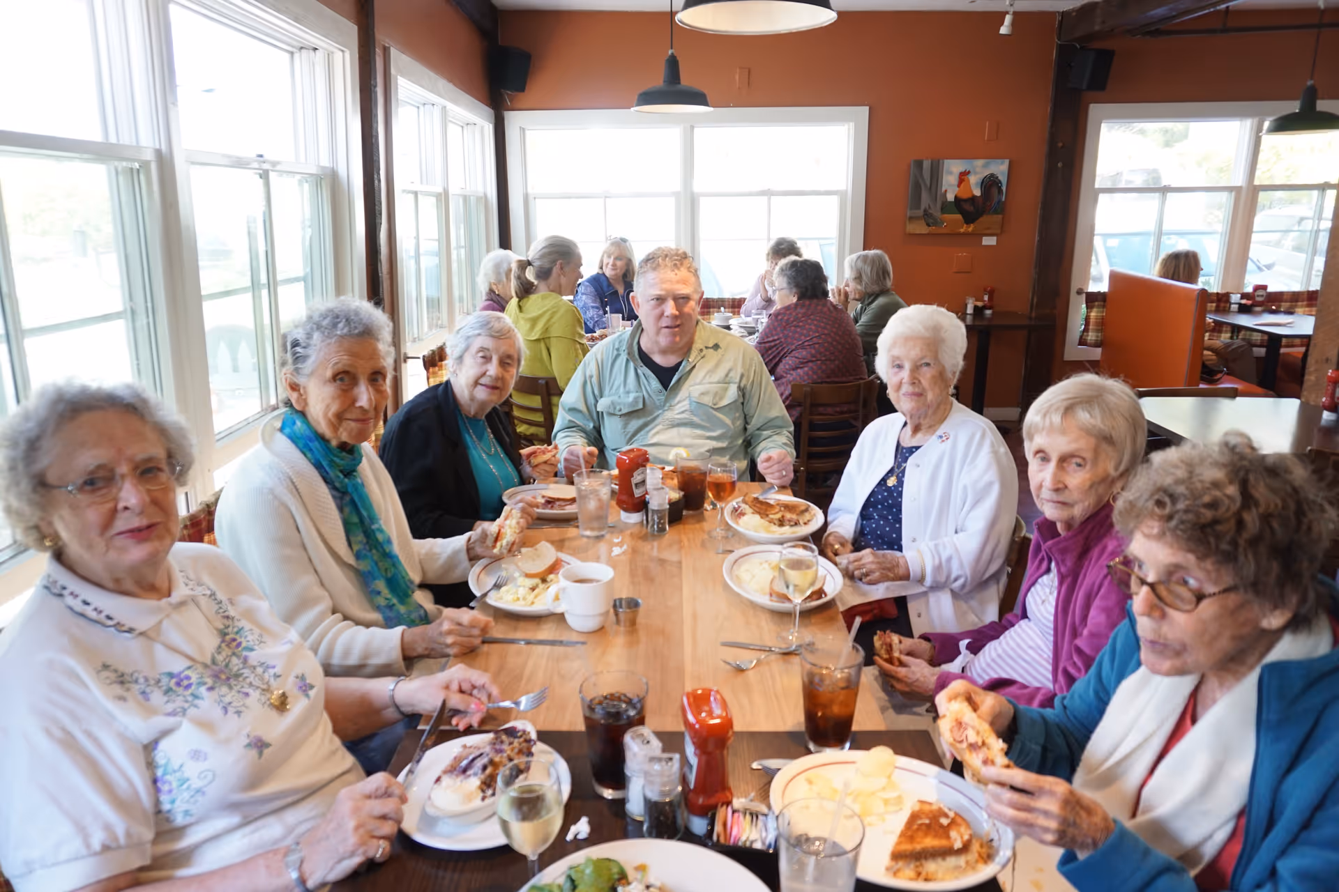 A group of elderly people and one middle-aged man sitting around a wooden dining table in a restaurant or dining room, enjoying a meal together. The table is set with plates of food, drinks, and condiments. Large windows let in natural light, and there are other diners visible in the background.