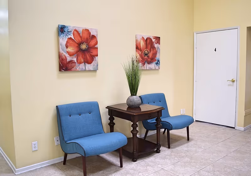 Indoor seating area with two blue chairs, a table, plant centerpiece, and floral art.