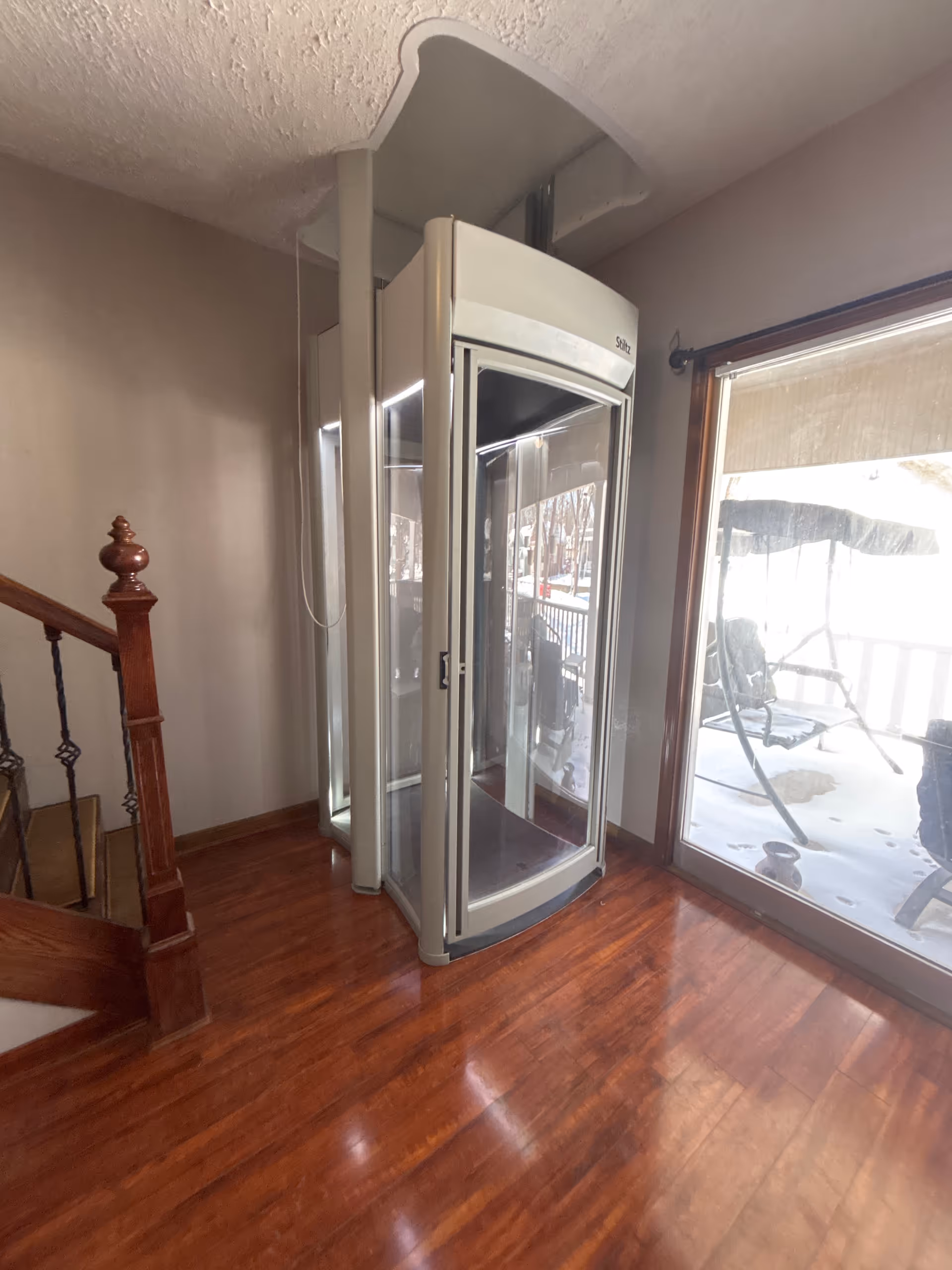 Indoor hallway with a compact glass-front residential elevator beside a wooden staircase.