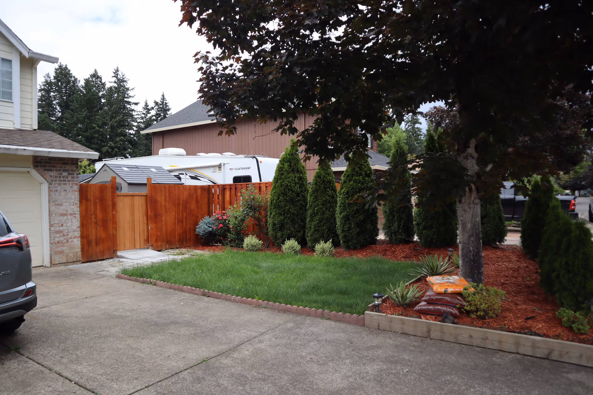 Residential exterior with a wooden fence, green lawn, mulch bed, and tall evergreens.