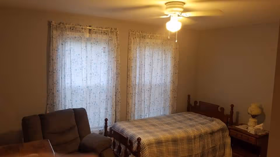 A cozy bedroom with a wooden bed, plaid quilt, nightstand, lamp, and sheer curtains.