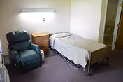 Small care-room with a single hospital-style bed, a wooden bedside table, a green recliner, and a wall-mounted light, with a doorway to a bathroom.