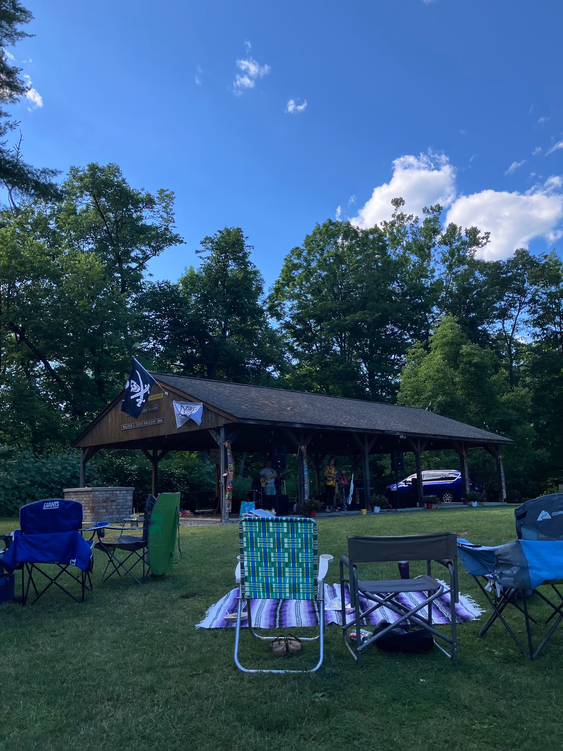 Outdoor pavilion gathering on grass with chairs, blankets, and people under trees.