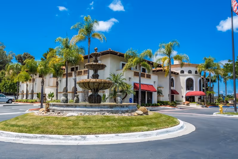 Exterior view of a senior living facility with a fountain, palm trees, and a circular driveway.