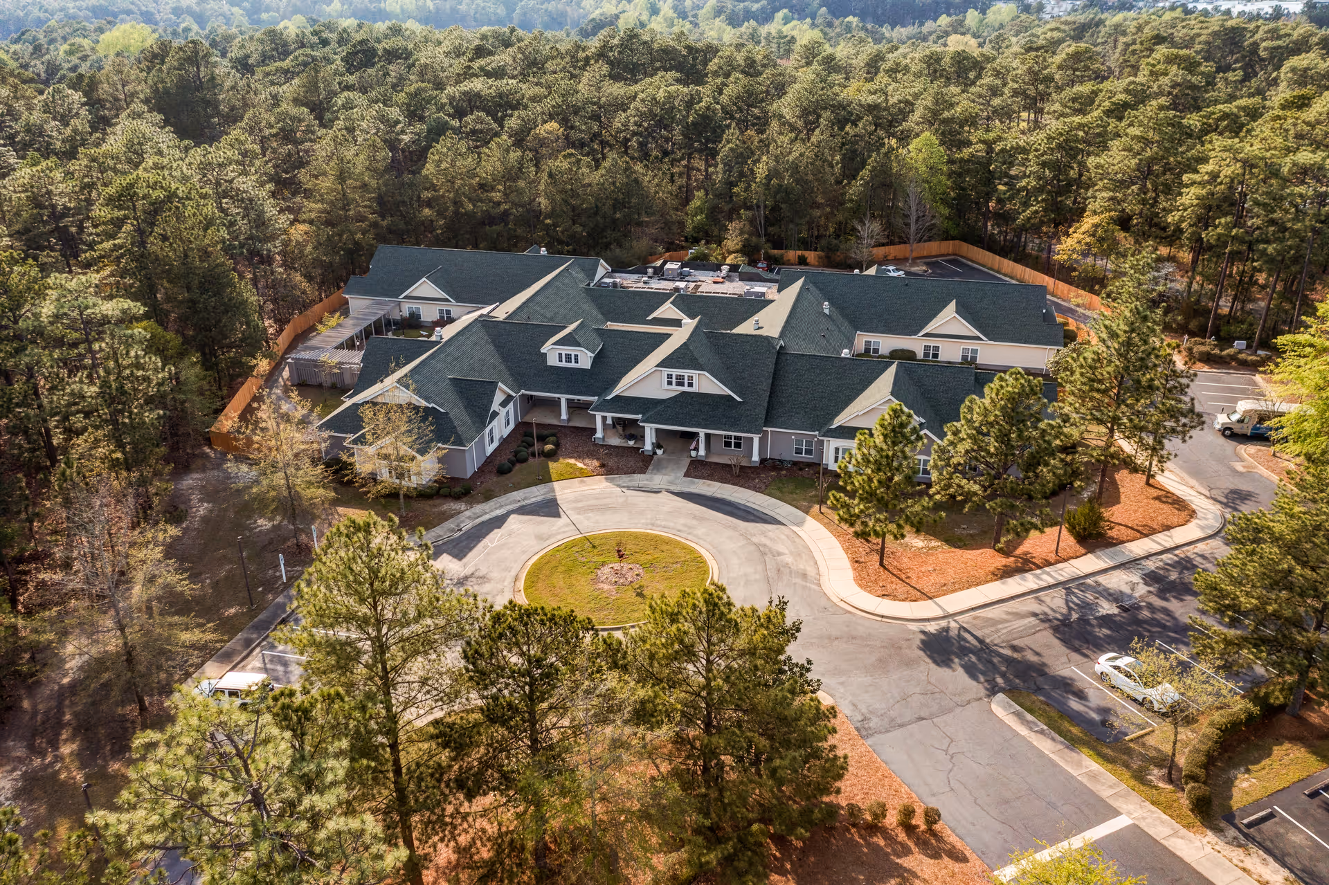 Aerial view of TerraBella Southern Pines facility surrounded by dense trees. The building has multiple peaked roofs with green shingles and a circular driveway in front. There are parking spaces and a few vehicles visible on the right side of the image.