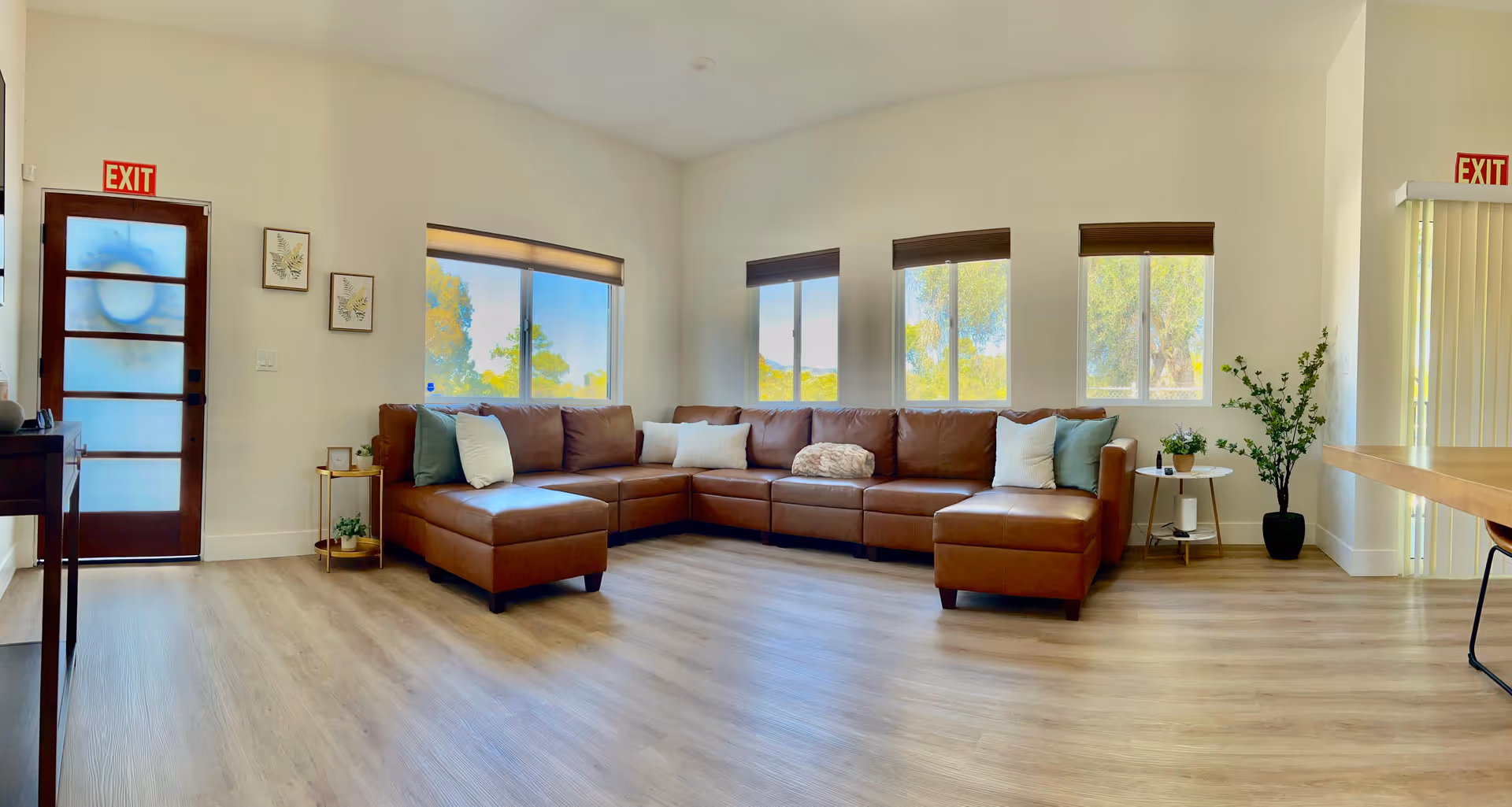 Spacious living room with brown sectional, light wood floor, windows, and potted plants.