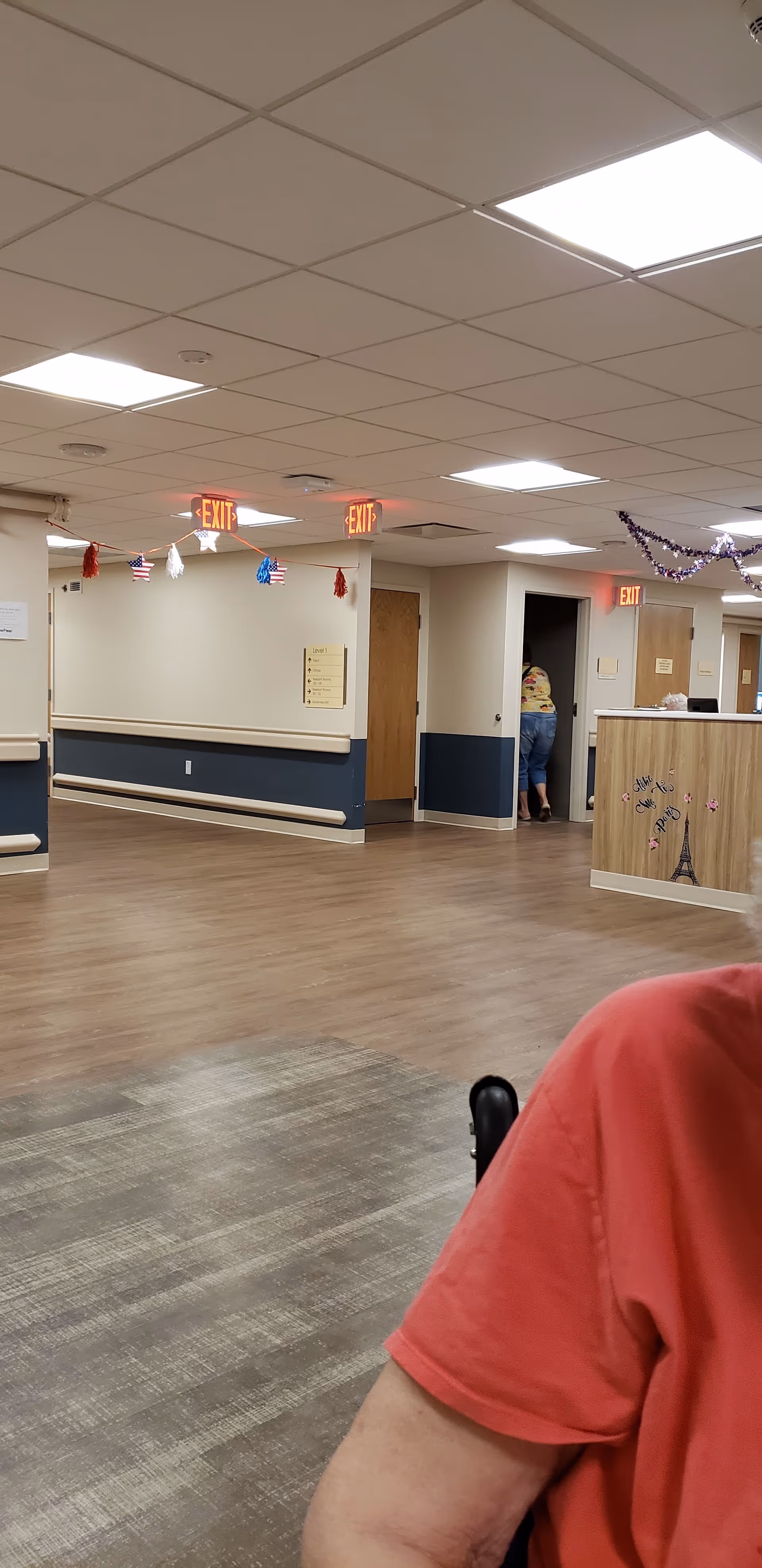 Interior hallway of a senior living facility with lighting, decorations, and a person near a doorway.