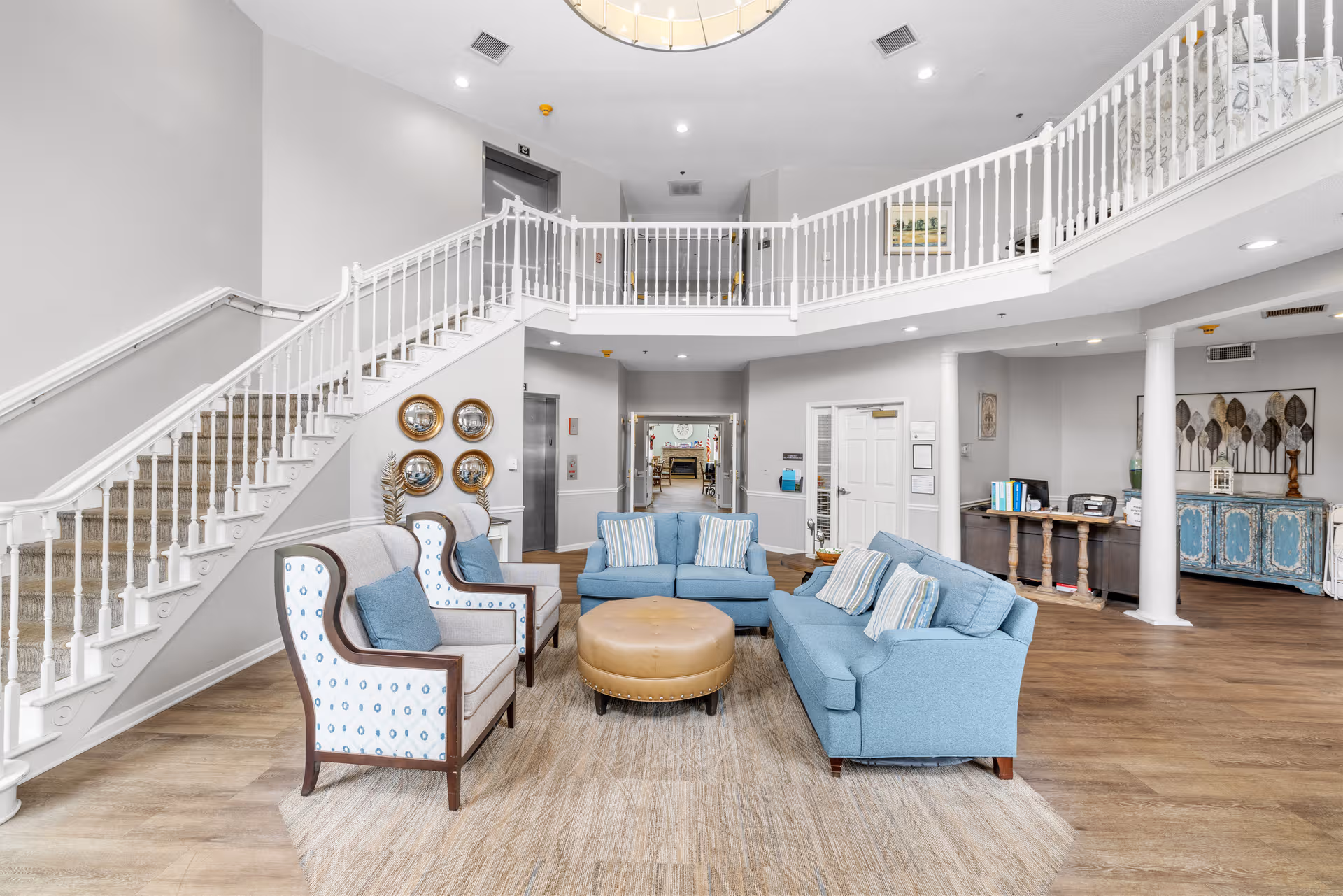 Bright senior living lobby with blue seating, staircase, and balcony railing in center.