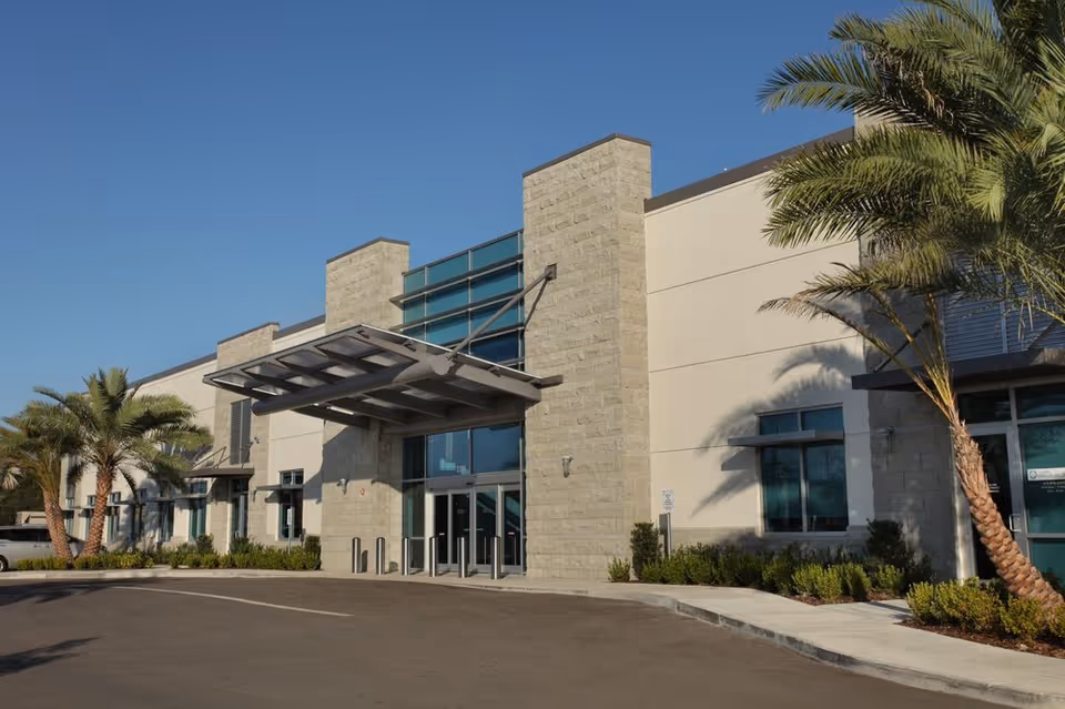 Modern building facade with glass entrance, stone columns, and palm trees on a sunny exterior.