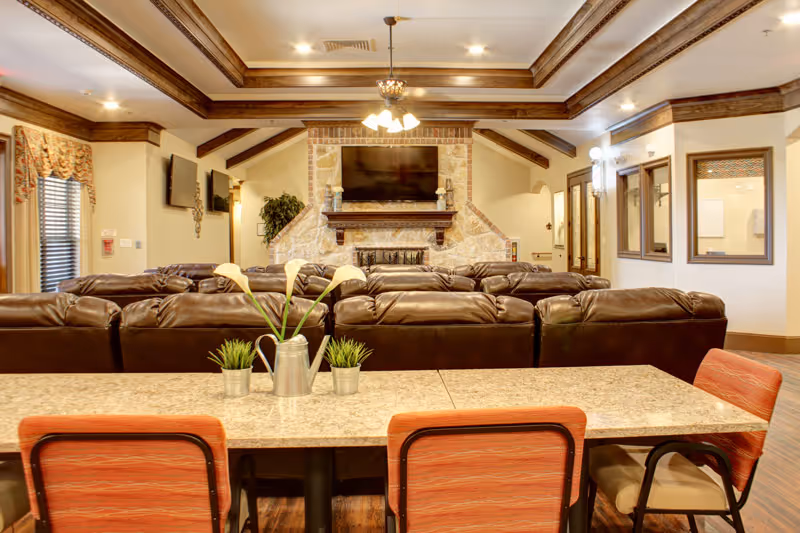 A cozy living room area with multiple brown leather sofas facing a stone fireplace with a mounted flat-screen TV above it. The room features a tray ceiling with wooden beams and a chandelier. In the foreground, there is a table with three orange cushioned chairs and small decorative plants on the table. The walls have windows and doors, and the floor is wooden.